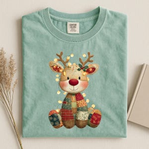 May include: A light teal Comfort Colors t-shirt featuring a cartoon reindeer with a red nose, patchwork body, and string lights. The reindeer has antlers and is decorated with holly. The shirt has a crew neck and a tag.