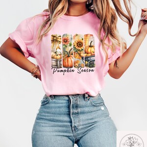May include: A light pink t-shirt featuring a fall-themed graphic with pumpkins, sunflowers, and the text "Pumpkin Season." The design includes pumpkins in a wooden crate, a sunflower, and a pumpkin in a wheelbarrow. The shirt is paired with blue jeans.