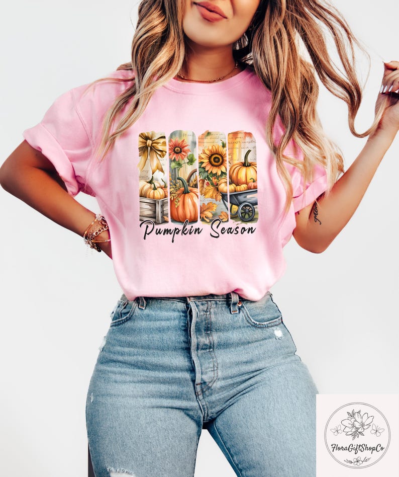 May include: A light pink t-shirt featuring a fall-themed graphic with pumpkins, sunflowers, and the text "Pumpkin Season." The design includes pumpkins in a wooden crate, a sunflower, and a pumpkin in a wheelbarrow. The shirt is paired with blue jeans.