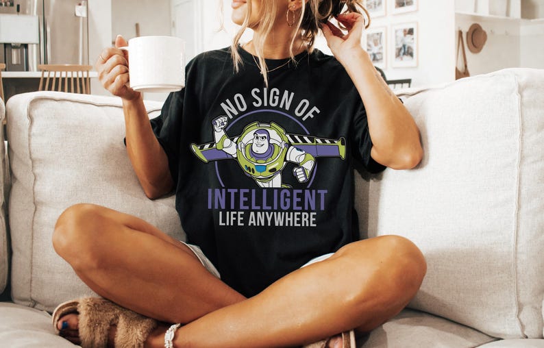 May include: Black t-shirt featuring a graphic of Buzz Lightyear with the text "NO SIGN OF INTELLIGENT LIFE ANYWHERE" in white and purple. The shirt is worn by a person sitting on a couch and holding a white mug.