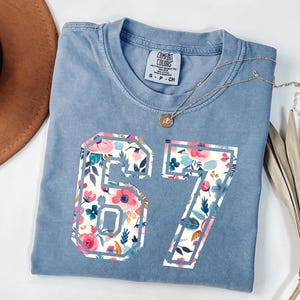 May include: A light blue t-shirt with the number "67" in a floral pattern. The shirt has a crew neck and a small tag on the upper left. A gold necklace with a pendant is draped across the shirt.