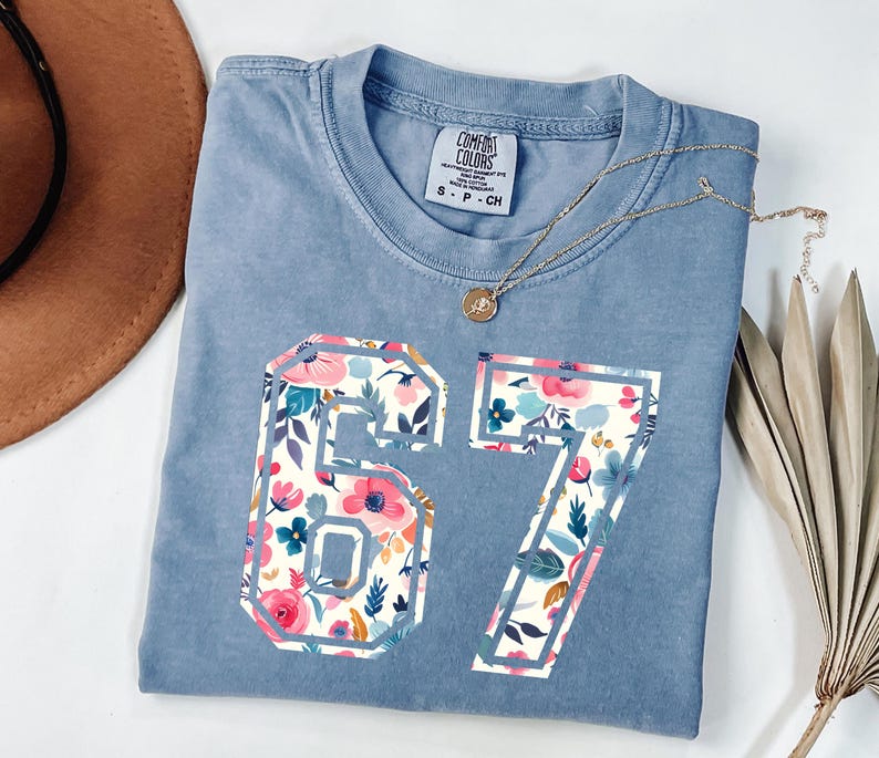 May include: A light blue t-shirt with the number "67" in a floral pattern. The shirt has a crew neck and a small tag on the upper left. A gold necklace with a pendant is draped across the shirt.