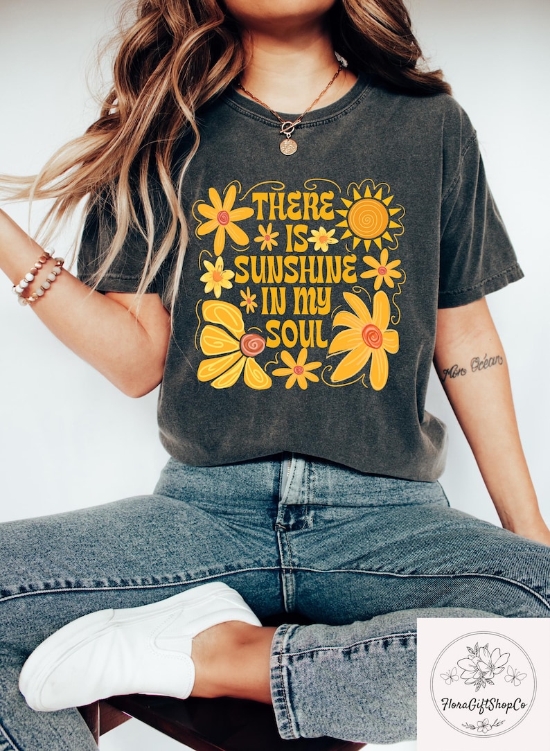 May include: Charcoal grey t-shirt featuring a retro floral design and the text "THERE IS SUNSHINE IN MY SOUL" in yellow. The shirt is worn with blue jeans and white trainers. The style has a positive, vintage aesthetic.