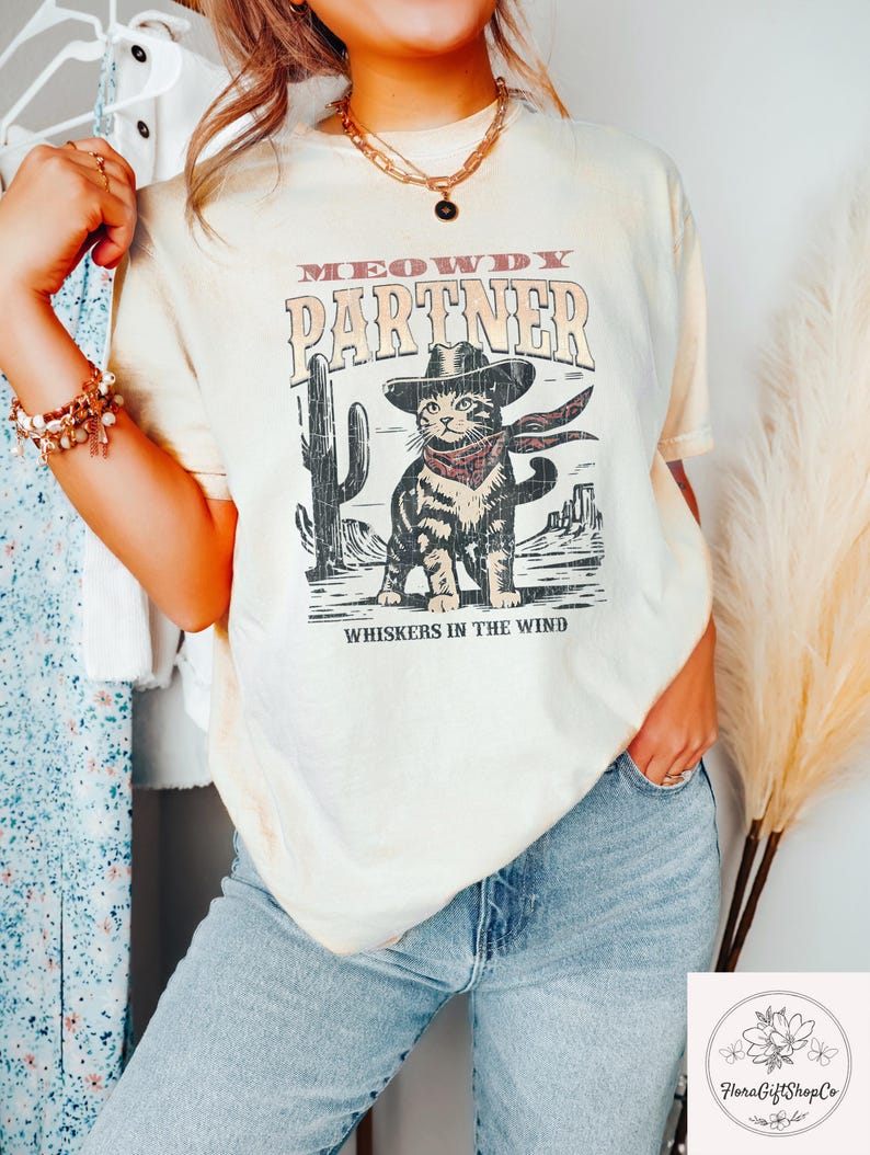 K&ouml;nnte beinhalten: Cremefarbenes T-Shirt mit einer Grafik einer Katze mit Cowboyhut und Halstuch, mit dem Text "MEOWDY PARTNER" und "WHISKERS IN THE WIND". Das Design beinhaltet einen Kaktus und eine W&uuml;stenlandschaft.