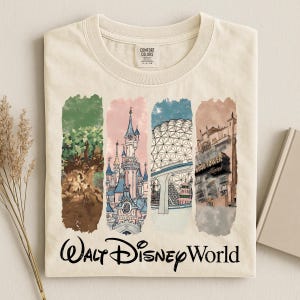 May include: Cream-colored t-shirt featuring a colorful illustration of Walt Disney World landmarks. The design includes images of a tree, a castle, a geodesic sphere, and a hotel, with "Walt Disney World" written below.
