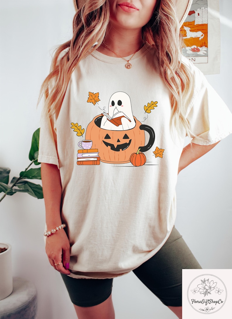 May include: Beige t-shirt featuring a Halloween-themed graphic. The design includes a ghost reading a book inside a pumpkin mug, surrounded by autumn leaves and a small pumpkin. A stack of books and a teacup are also present.