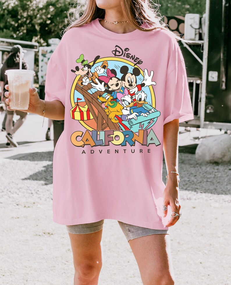 May include: A light pink t-shirt featuring a Disney graphic with Mickey Mouse, Minnie Mouse, Donald Duck, Goofy, and other characters on a roller coaster. The text "CALIFORNIA ADVENTURE" is below the graphic.