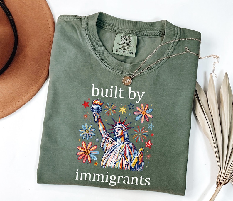 May include: An olive green t-shirt featuring a graphic of the Statue of Liberty surrounded by colorful flowers and stars. The text "built by immigrants" is printed in white above and below the graphic. The shirt has a crew neck and short sleeves.