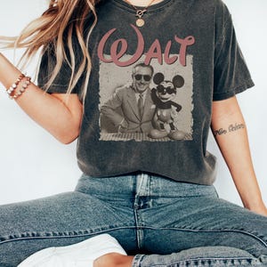 May include: Dark gray t-shirt featuring a vintage-style graphic of Walt Disney and Mickey Mouse. The word "Walt" is printed in a pink, retro font above the image. The shirt is a casual wear item.