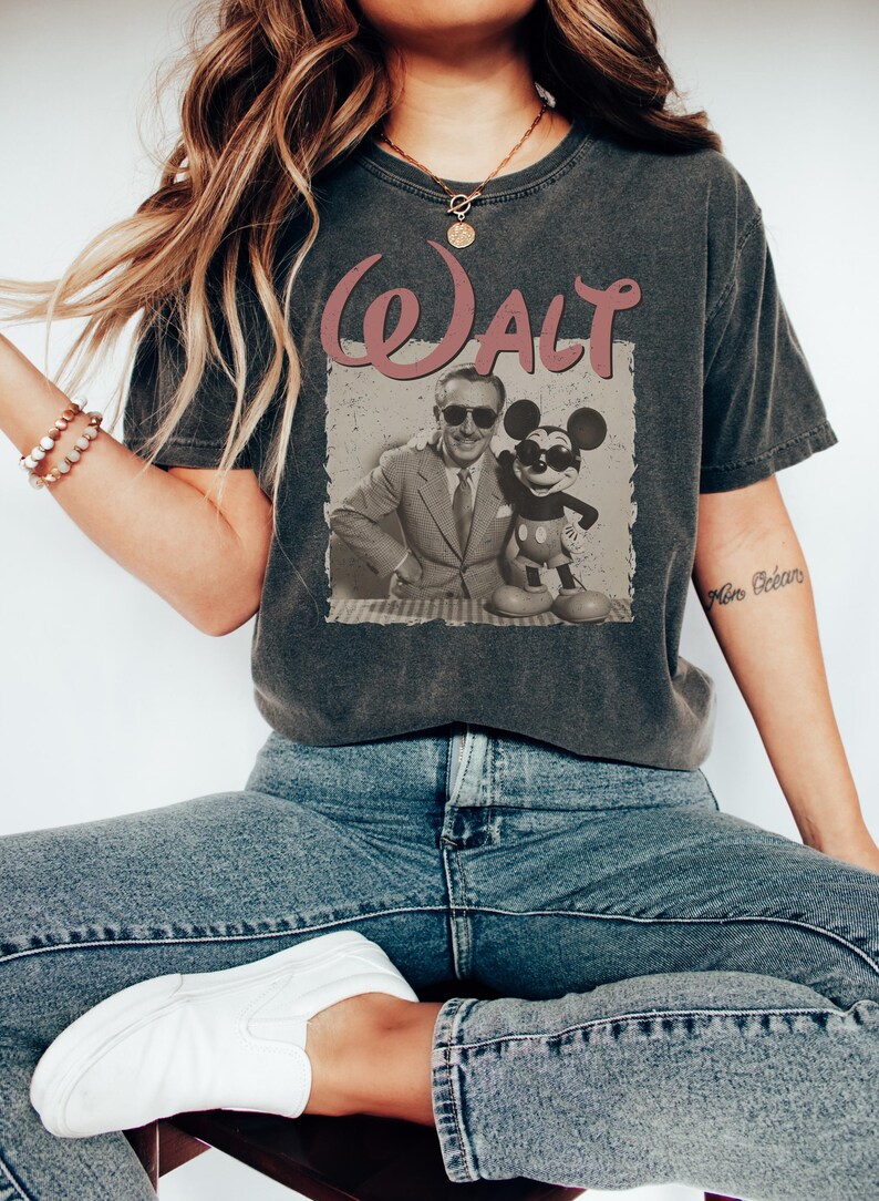 May include: Dark gray t-shirt featuring a vintage-style graphic of Walt Disney and Mickey Mouse. The word "Walt" is printed in a pink, retro font above the image. The shirt is a casual wear item.