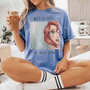 May include: A periwinkle blue short-sleeve t-shirt featuring a square graphic print of a red-haired character with large eyes. The text "BUT DADDY" is printed above the graphic and "I Love Him" is printed below the graphic in a black script font.