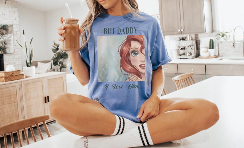 May include: A periwinkle blue short-sleeve t-shirt featuring a square graphic print of a red-haired character with large eyes. The text "BUT DADDY" is printed above the graphic and "I Love Him" is printed below the graphic in a black script font.