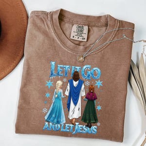 May include: Brown t-shirt with the text "LET IT GO AND LET JESUS" and a graphic of Jesus with two cartoon characters. The shirt has a crew neck and short sleeves. A silver necklace with a pendant is on the shirt.