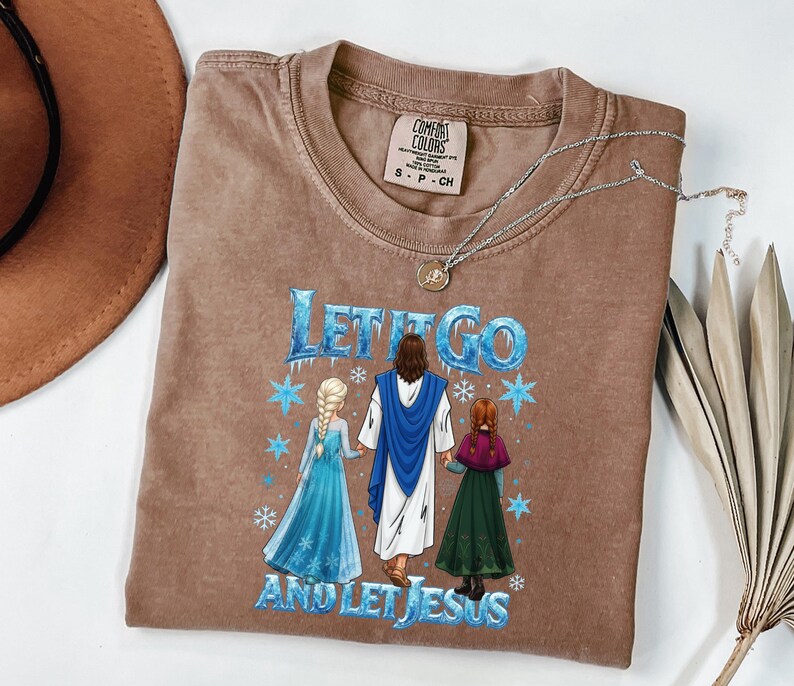 May include: Brown t-shirt with the text "LET IT GO AND LET JESUS" and a graphic of Jesus with two cartoon characters. The shirt has a crew neck and short sleeves. A silver necklace with a pendant is on the shirt.