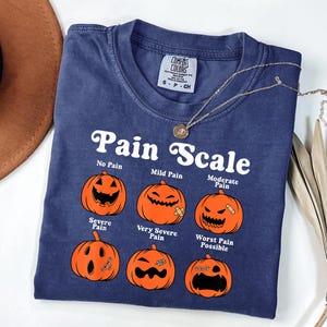 May include: A navy blue t-shirt with the text "Pain Scale" and illustrations of jack-o'-lanterns depicting different levels of pain. The shirt includes a small tag and a silver necklace. The shirt is a Comfort Colors brand.