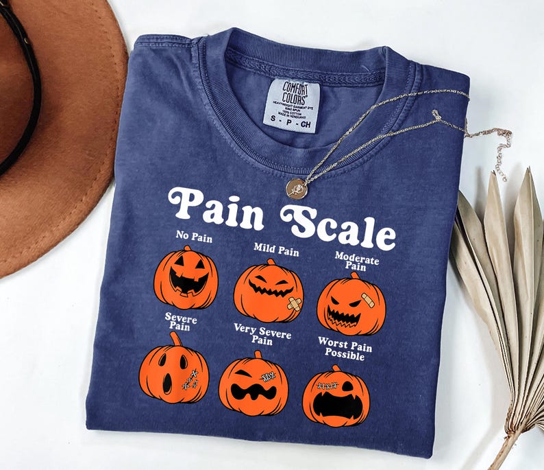 May include: A navy blue t-shirt with the text "Pain Scale" and illustrations of jack-o'-lanterns depicting different levels of pain. The shirt includes a small tag and a silver necklace. The shirt is a Comfort Colors brand.