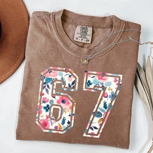 May include: A brown Comfort Colors t-shirt with the number 67 in a floral design. The floral pattern includes pink, blue, and white flowers. A silver necklace with a pendant is draped across the shirt.