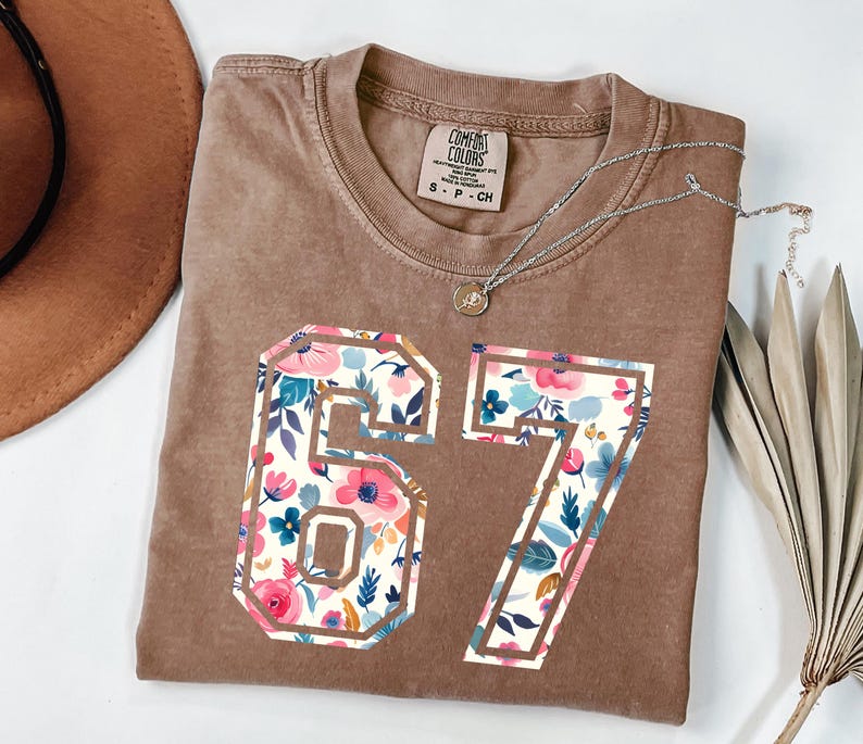 May include: A brown Comfort Colors t-shirt with the number 67 in a floral design. The floral pattern includes pink, blue, and white flowers. A silver necklace with a pendant is draped across the shirt.