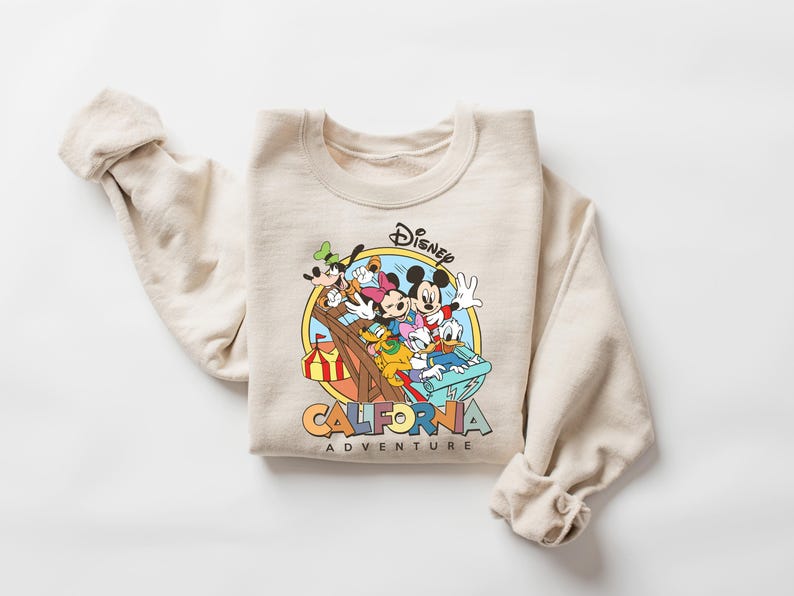 May include: Beige crewneck sweatshirt featuring a colorful graphic of Mickey Mouse, Minnie Mouse, Donald Duck, Goofy, and Pluto. The graphic is set within a yellow circle and includes the text "Disney California Adventure".