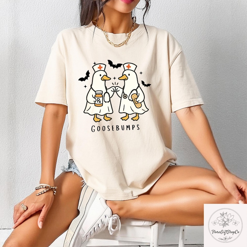 May include: Cream-colored t-shirt featuring a cartoon design of two ducks dressed as nurses, with bats and the word "GOOSEBUMPS". The ducks are holding medical items. The shirt is a casual, short-sleeved style.
