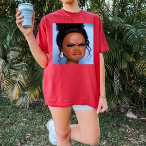 May include: Red t-shirt with a square graphic of a doll's face with a displeased expression. The doll has dark hair, makeup, and large earrings. The person is holding a blue collapsible cup. The background is green.