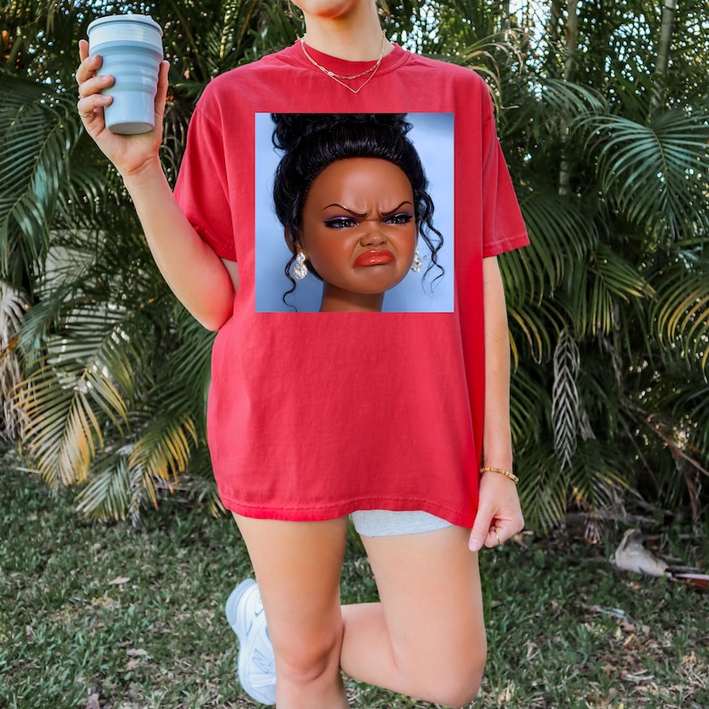 May include: Red t-shirt with a square graphic of a doll's face with a displeased expression. The doll has dark hair, makeup, and large earrings. The person is holding a blue collapsible cup. The background is green.
