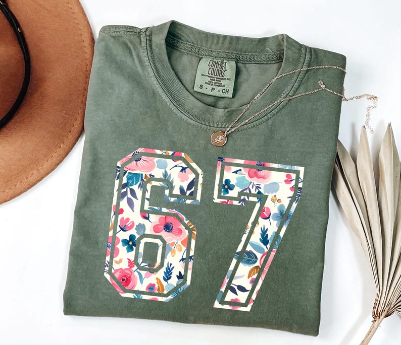 May include: Olive green t-shirt with the numbers 67 in a floral pattern. The numbers are outlined in a white border. A gold necklace with a pendant is on the shirt.