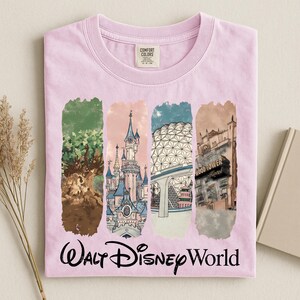 May include: A light pink t-shirt featuring four vertical rectangular panels with illustrations of Walt Disney World landmarks. The shirt has the words "Walt Disney World" in a cursive font at the bottom.