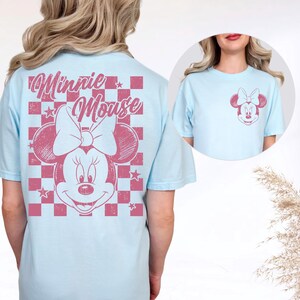 May include: Pale blue t-shirt with a pink Minnie Mouse design on the back and a smaller version on the front. The back graphic includes the words "Minnie Mouse" and a chequered pattern. The shirt has short sleeves and a round neck.