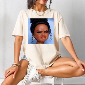 May include: Beige t-shirt featuring a graphic of a doll's face with a displeased expression. The doll has dark skin, black hair, and red lipstick. The t-shirt is a neutral color and has short sleeves.