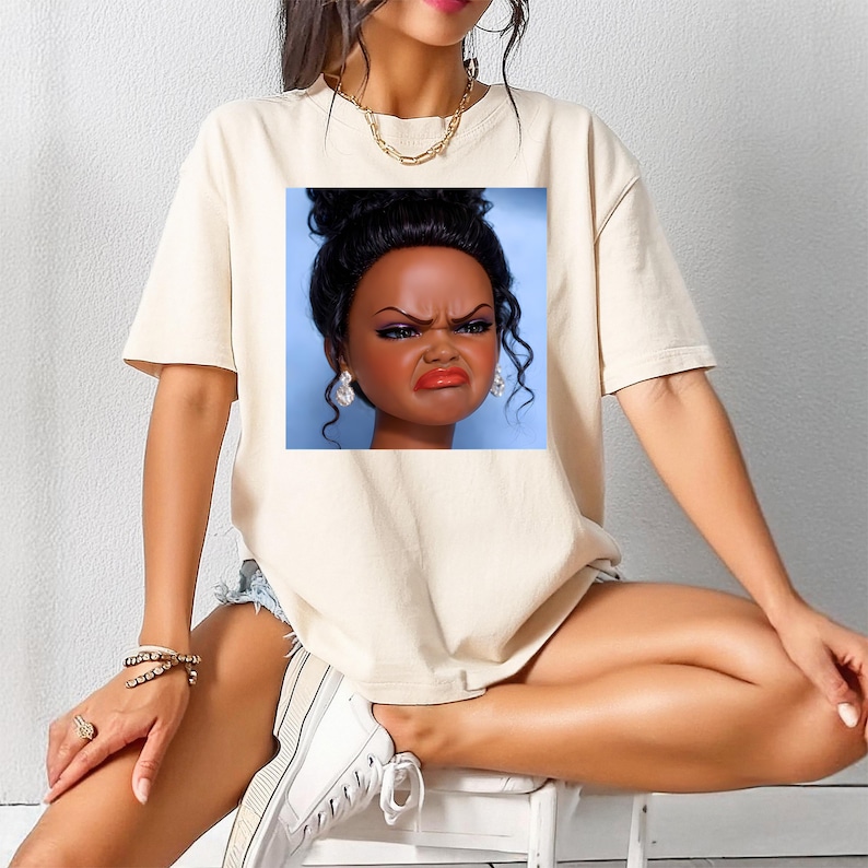 May include: Beige t-shirt featuring a graphic of a doll's face with a displeased expression. The doll has dark skin, black hair, and red lipstick. The t-shirt is a neutral color and has short sleeves.