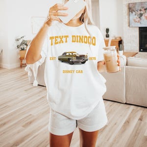 Peut inclure: T-shirt blanc avec un motif de voiture verte et le texte "TEXT DINOCO EST 1975 DISNEY CAR" en or. Le t-shirt est de coupe ample et d&eacute;contract&eacute;e.