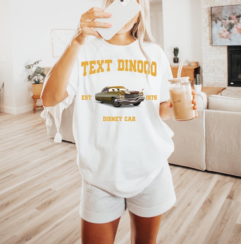 Peut inclure: T-shirt blanc avec un motif de voiture verte et le texte "TEXT DINOCO EST 1975 DISNEY CAR" en or. Le t-shirt est de coupe ample et d&eacute;contract&eacute;e.