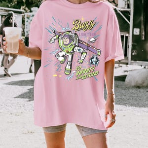 May include: A light pink t-shirt featuring a graphic of Buzz Lightyear from Toy Story. The design includes the text "Buzzy Lightyear" and "To Infinity and Beyond" in a playful font, with a space-themed background.