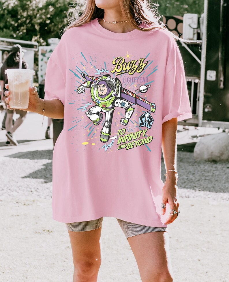 May include: A light pink t-shirt featuring a graphic of Buzz Lightyear from Toy Story. The design includes the text "Buzzy Lightyear" and "To Infinity and Beyond" in a playful font, with a space-themed background.