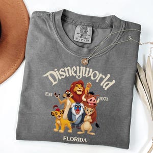 Pode incluir: Uma t-shirt cinzento-carv&atilde;o de gola redonda com um gr&aacute;fico das personagens da Disney World, incluindo Simba, Nala, Timon, Pumbaa, Mufasa e Rafiki. O texto diz Disneyworld Est 1971 Florida. A t-shirt tem uma etiqueta da marca Comfort Colors.