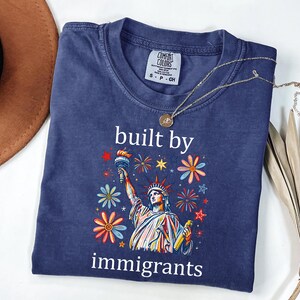May include: A navy blue short-sleeve t-shirt featuring a graphic of the Statue of Liberty surrounded by colorful flowers and fireworks. The text "built by immigrants" is printed in white above and below the graphic. A gold necklace rests on the collar.