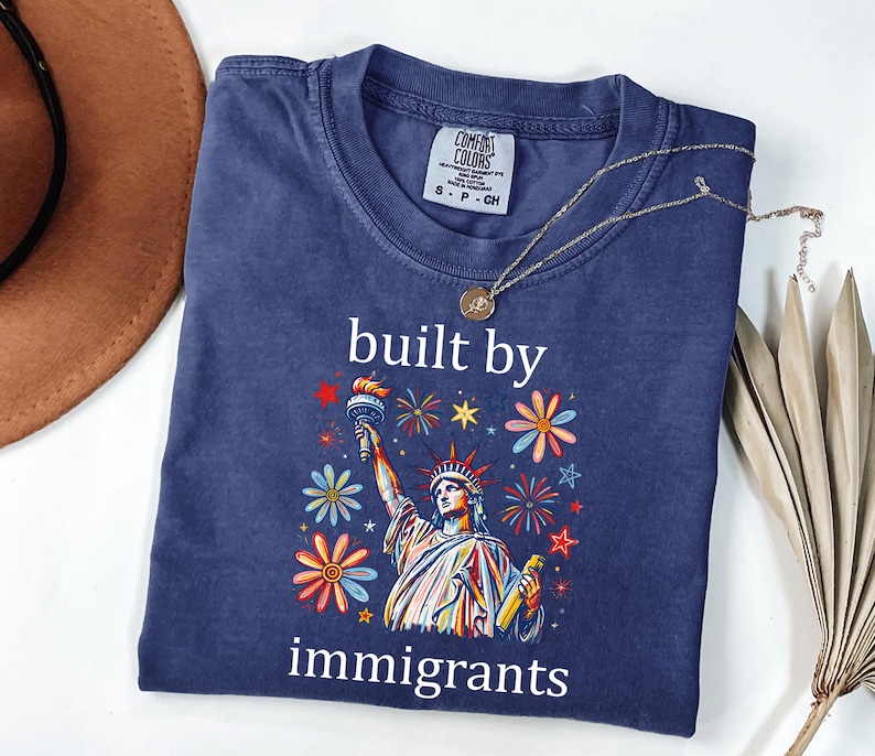 May include: A navy blue short-sleeve t-shirt featuring a graphic of the Statue of Liberty surrounded by colorful flowers and fireworks. The text "built by immigrants" is printed in white above and below the graphic. A gold necklace rests on the collar.
