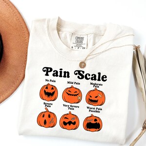 May include: Cream-colored t-shirt with a "Pain Scale" graphic featuring orange jack-o'-lanterns representing different levels of pain. The text includes "No Pain" to "Worst Pain Possible". A gold necklace is draped over the shirt.