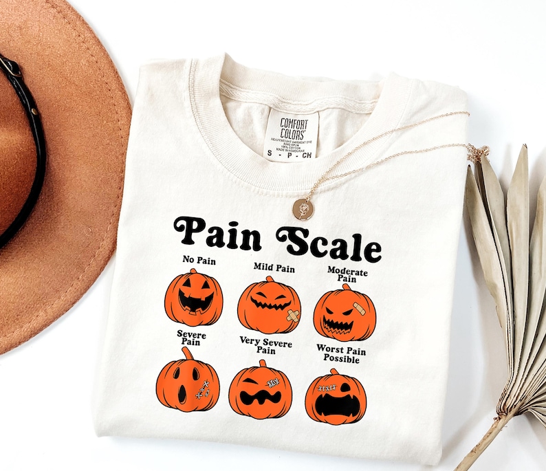 May include: Cream-colored t-shirt with a "Pain Scale" graphic featuring orange jack-o'-lanterns representing different levels of pain. The text includes "No Pain" to "Worst Pain Possible". A gold necklace is draped over the shirt.