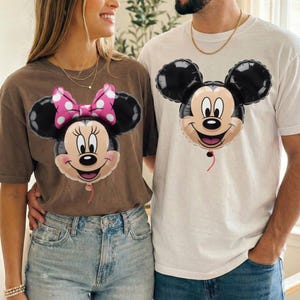 Peut inclure: Un t-shirt marron avec un motif de ballon Minnie Mouse portant un n&oelig;ud rose &agrave; pois et un t-shirt blanc avec un motif de ballon Mickey Mouse. Les deux t-shirts pr&eacute;sentent les visages des personnages sous forme d'illustrations de ballons gonfl&eacute;s.