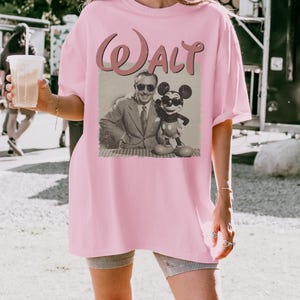 May include: A light pink t-shirt featuring a vintage-style graphic of Walt Disney with Mickey Mouse. The word "Walt" is printed above the image in a cursive font. The shirt is a loose fit.