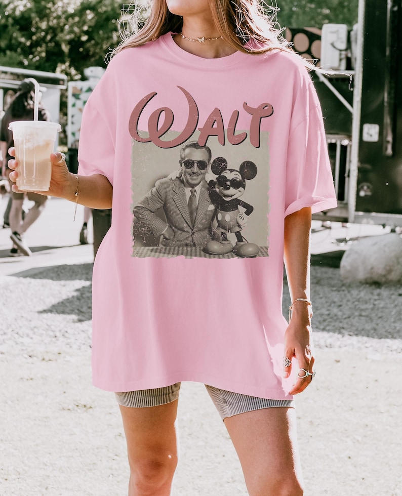 May include: A light pink t-shirt featuring a vintage-style graphic of Walt Disney with Mickey Mouse. The word "Walt" is printed above the image in a cursive font. The shirt is a loose fit.