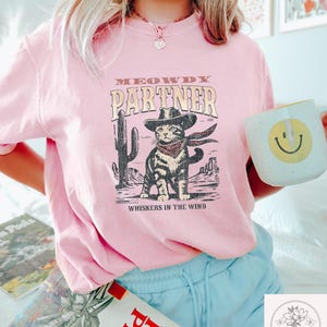 K&ouml;nnte beinhalten: Hellrosa T-Shirt mit einem Cartoon einer Katze mit Cowboyhut und Halstuch, mit dem Text "MEOWDY PARTNER" und "WHISKERS IN THE WIND". Das Design zeigt einen Kaktus und eine W&uuml;stenlandschaft.