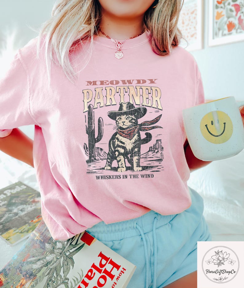 K&ouml;nnte beinhalten: Hellrosa T-Shirt mit einem Cartoon einer Katze mit Cowboyhut und Halstuch, mit dem Text "MEOWDY PARTNER" und "WHISKERS IN THE WIND". Das Design zeigt einen Kaktus und eine W&uuml;stenlandschaft.
