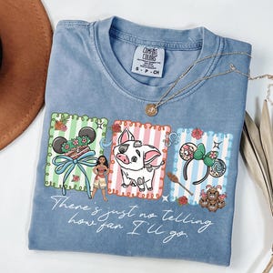 May include: A blue short-sleeved t-shirt featuring a graphic print of Moana, Pua the pig, and two sets of mouse ears in scalloped frames. The text "There's just no telling how far I'll go" is printed below the characters in a white script font.
