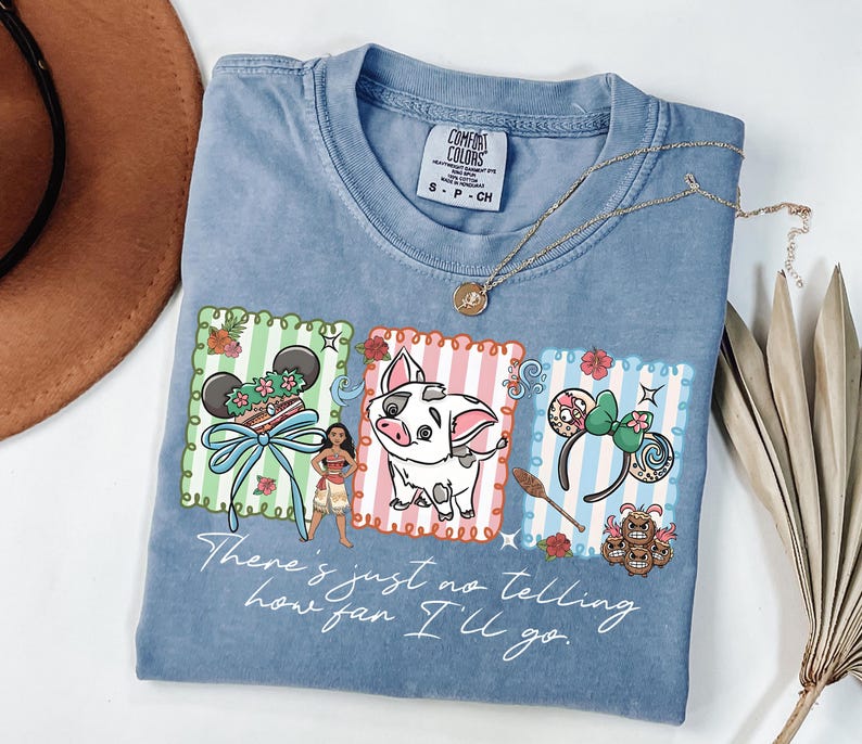 May include: A blue short-sleeved t-shirt featuring a graphic print of Moana, Pua the pig, and two sets of mouse ears in scalloped frames. The text "There's just no telling how far I'll go" is printed below the characters in a white script font.