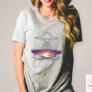 Op de afbeelding: Grijs t-shirt met een afbeelding van een UFO boven een zonsondergang. Het ontwerp toont het ruimtevaartuig en het landschap eronder, met paarse, oranje en grijze kleuren. Het t-shirt is gemaakt van zacht materiaal.