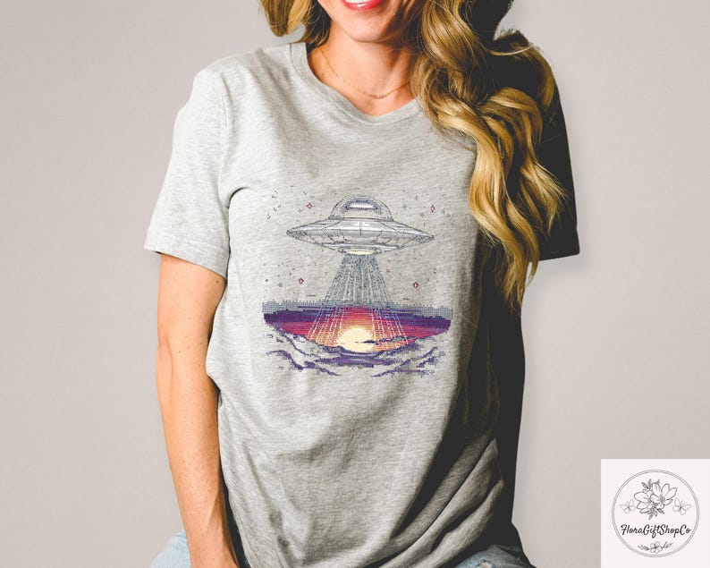 Op de afbeelding: Grijs t-shirt met een afbeelding van een UFO boven een zonsondergang. Het ontwerp toont het ruimtevaartuig en het landschap eronder, met paarse, oranje en grijze kleuren. Het t-shirt is gemaakt van zacht materiaal.