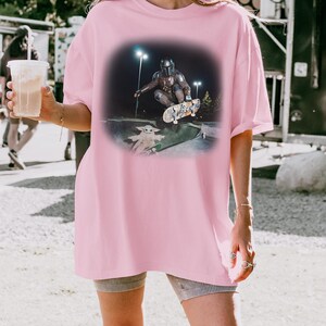 Op de afbeelding: Roze oversized T-shirt met een afbeelding van een personage met helm die in een skatepark skatet met een kleine groene alien. De afbeelding staat tegen een donkere achtergrond met heldere lichten.